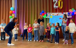 Kinder des Montessori Kinderhauses stehen bei der FunFace-Geburtstagsfeier auf der Bühne; Andrea Schreiber steht mit Mikrofon davor.