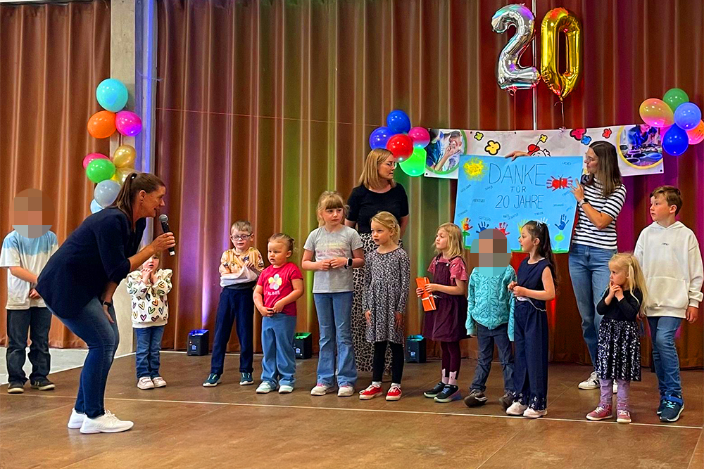 Kinder des Montessori Kinderhauses stehen bei der FunFace-Geburtstagsfeier auf der Bühne; Andrea Schreiber steht mit Mikrofon davor.